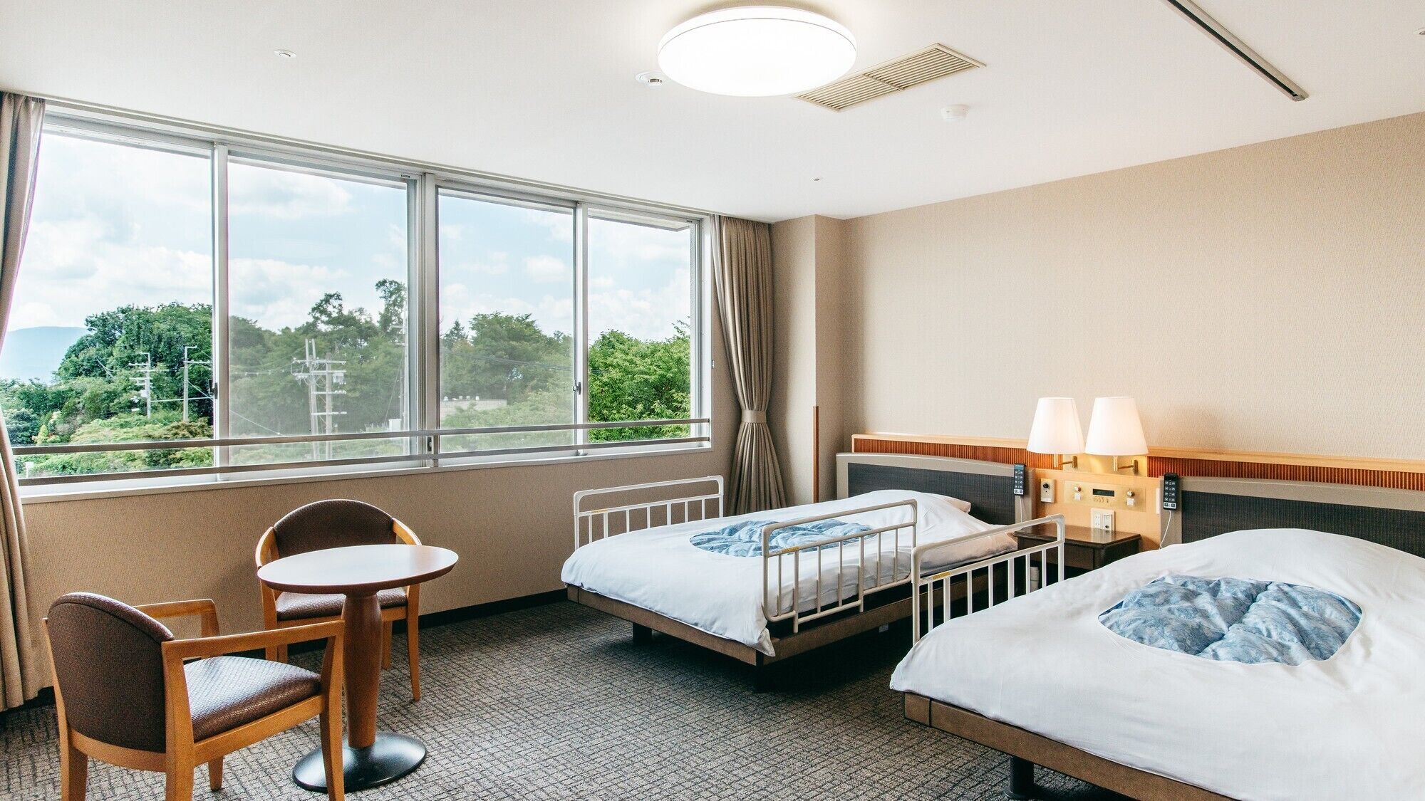 A hotel room with two beds, a small table, a chair, a window with a view of greenery, and a ceiling light.