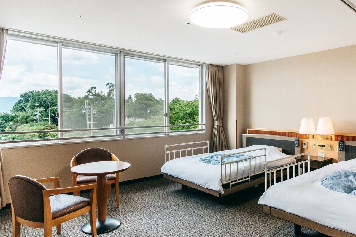 Une chambre d’hôtel avec deux lits, une petite table, une chaise, une fenêtre donnant sur un espace verdoyant et un luminaire au plafond.