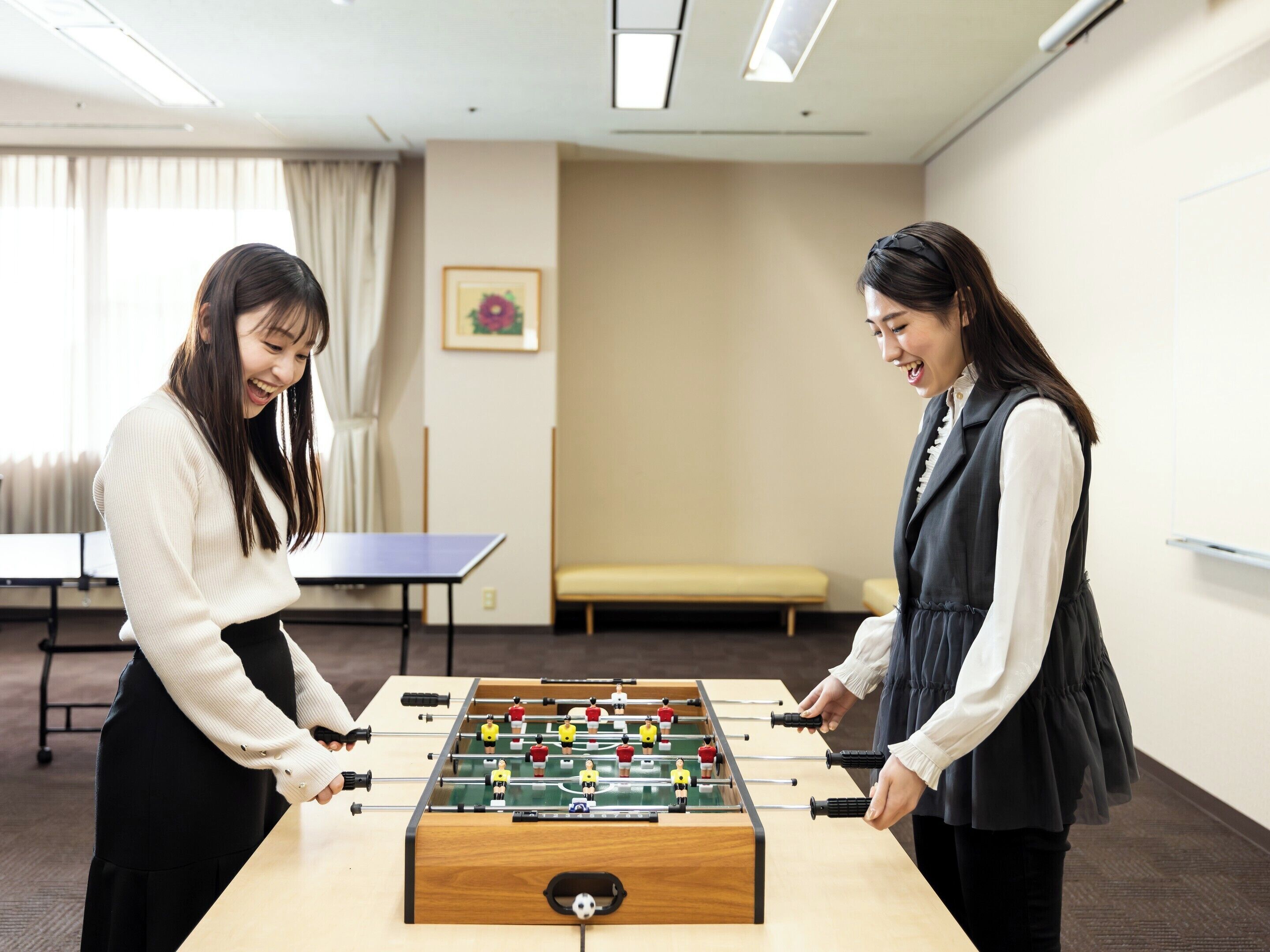 Duas pessoas jogando futebol de mesa em um espaço com mesa de pingue-pongue, um sofá e um quadro na parede.