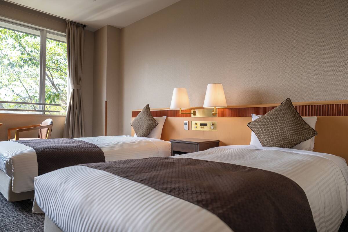 Une chambre d’hôtel avec deux lits, une table de chevet, une lampe et une vue sur les arbres par la fenêtre.