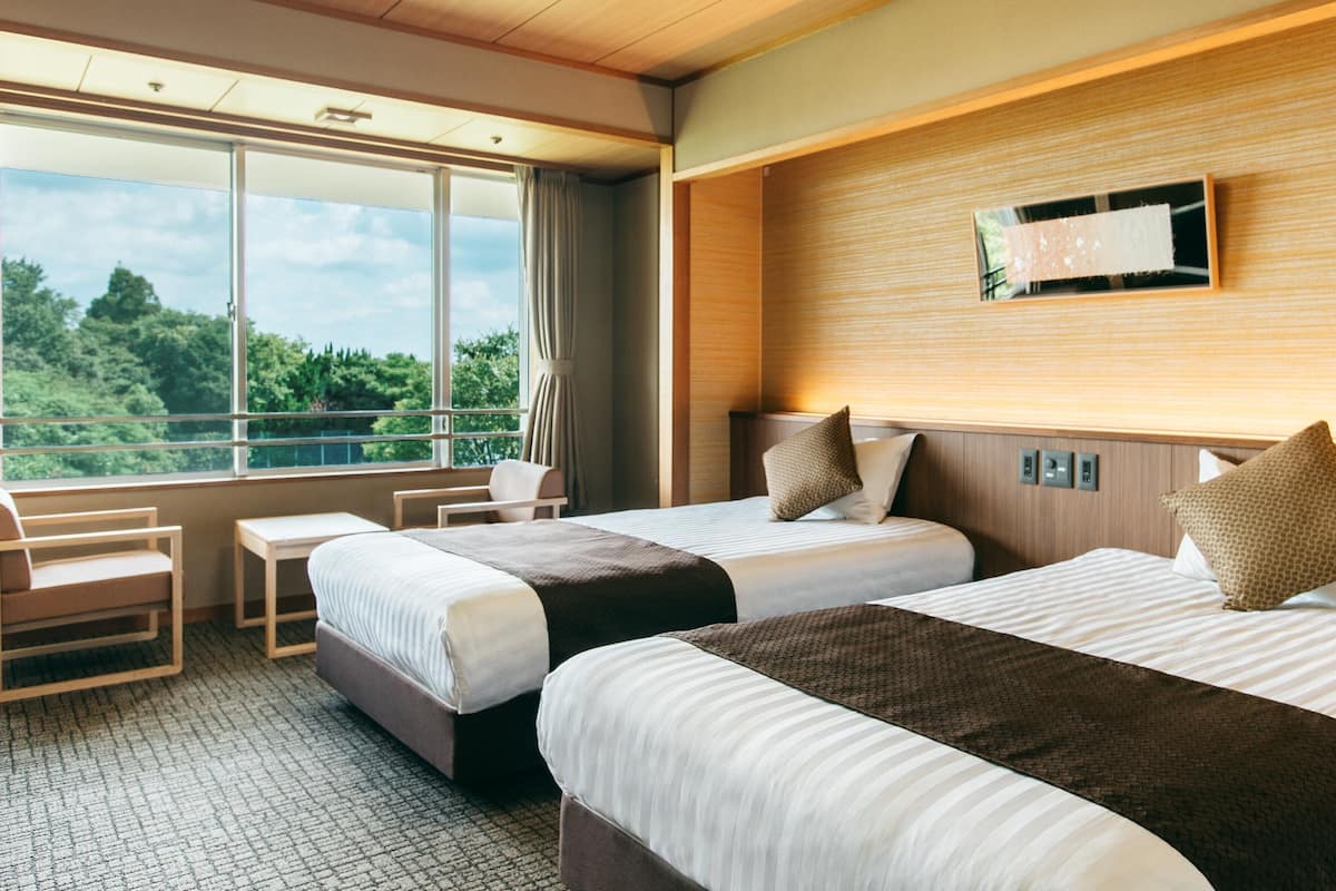 Une chambre d’hôtel avec deux lits, un bureau et une grande fenêtre donnant sur un paysage verdoyant.