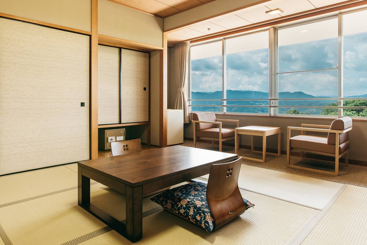 Une pièce de style japonais traditionnel, avec un sol recouvert de tatamis, une basse table en bois, deux chaises et une vue sur les montagnes par de grandes fenêtres.