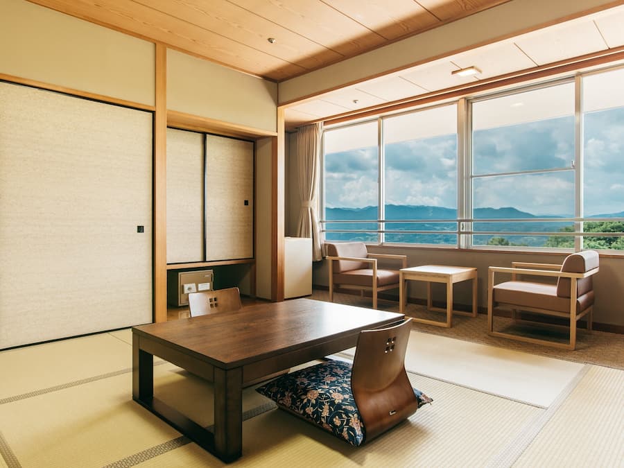 A traditional Japanese room with tatami flooring, a low wooden table, two chairs, and a view of mountains through large windows.