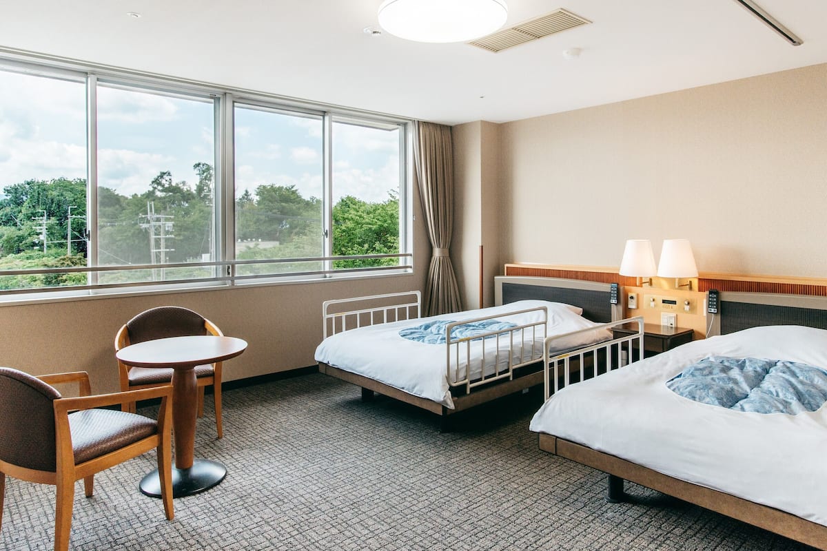 Une chambre d’hôtel avec deux lits, un bureau et une chaise. Une grande fenêtre offre une vue sur la verdure environnante.