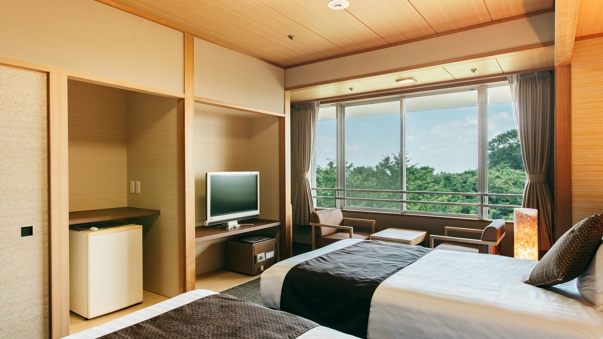 A hotel room with a bed, a desk, a television, and a view of greenery through large windows.