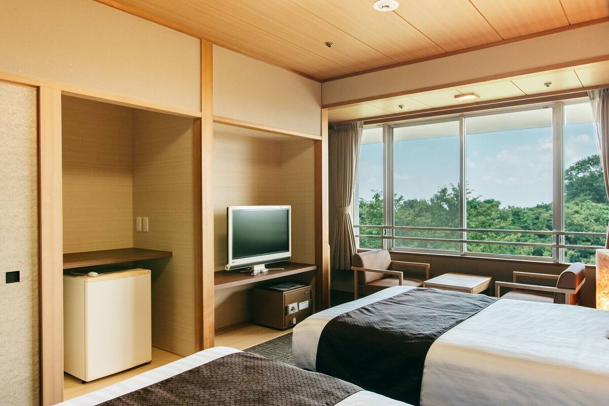 Une chambre d’hôtel équipée d’un lit, d’un bureau, d’une télévision et offrant une vue sur la verdure grâce à de grandes fenêtres.