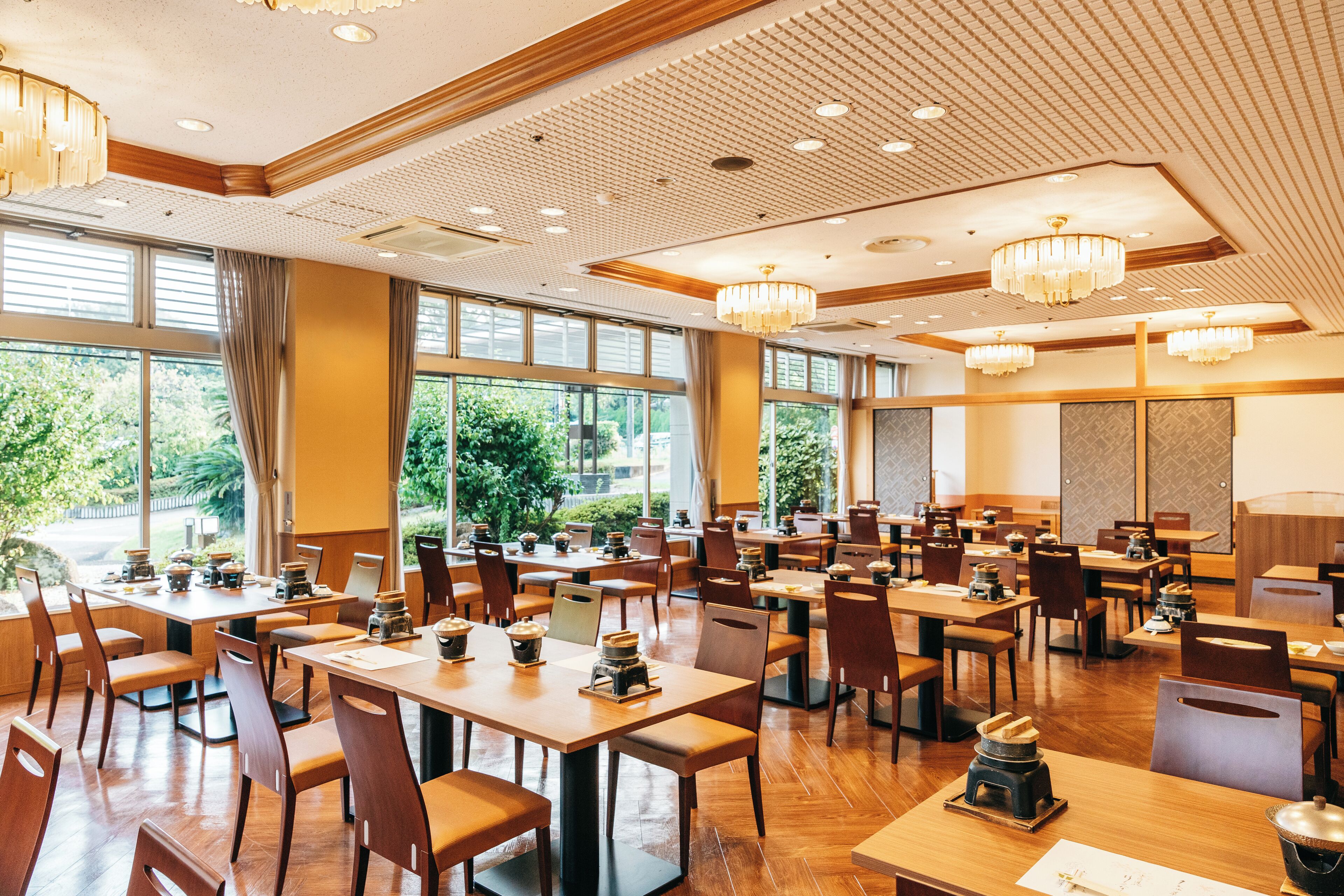 A spacious restaurant with multiple tables and chairs, large windows offering a view of greenery outside, and elegant chandeliers.