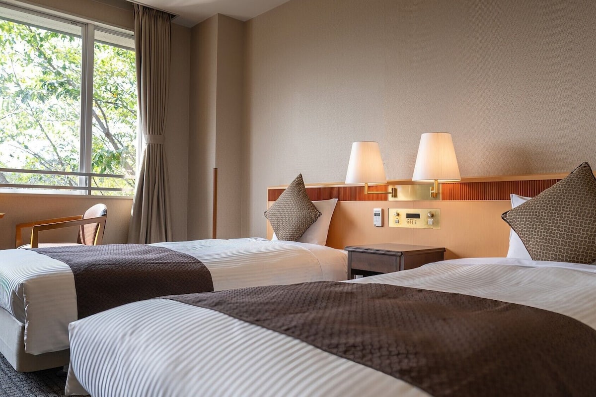 Une chambre d’hôtel avec deux lits, une table de chevet, un bureau et une fenêtre avec des rideaux.