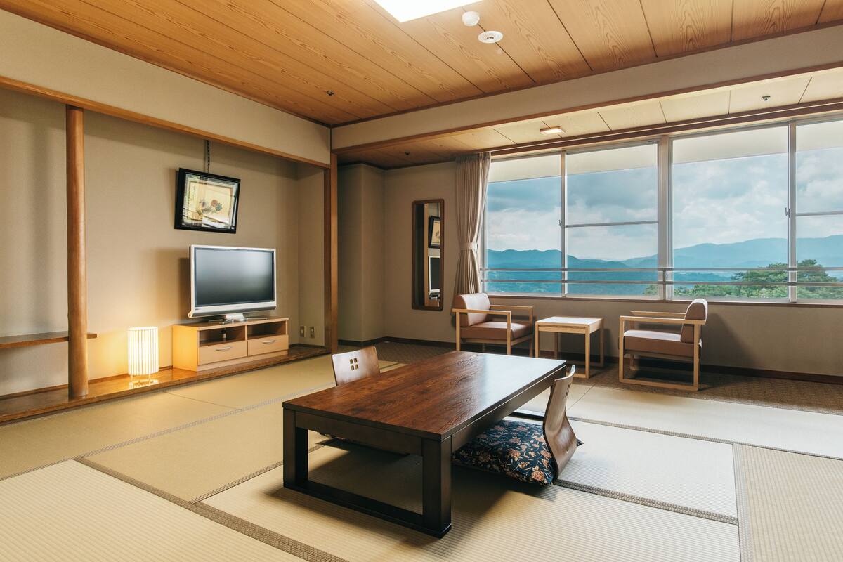 Une pièce de style japonais traditionnel, avec un sol recouvert de tatamis, une table en bois, un téléviseur et une grande fenêtre donnant sur la montagne.