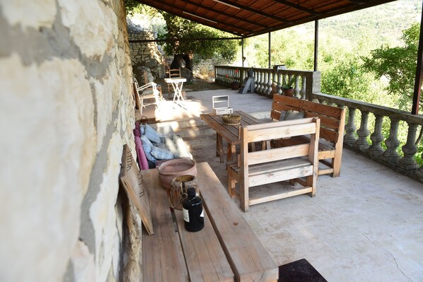 A covered outdoor seating area with stone walls, wooden furniture, and a view of greenery.