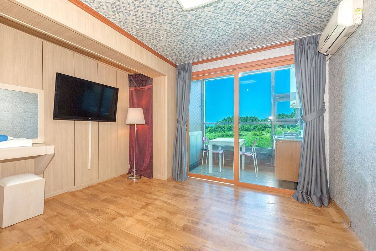 Habitación de hotel moderna con piso de madera, televisor de pantalla plana, un mueble blanco pequeño, una lámpara y un ventanal corredizo que da a un balcón con vista a zonas verdes.