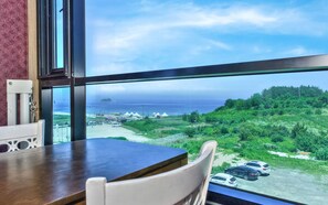A room with a large window offering a view of a beach, parked cars, and a distant island. - Fairy Pension (Sokcho)