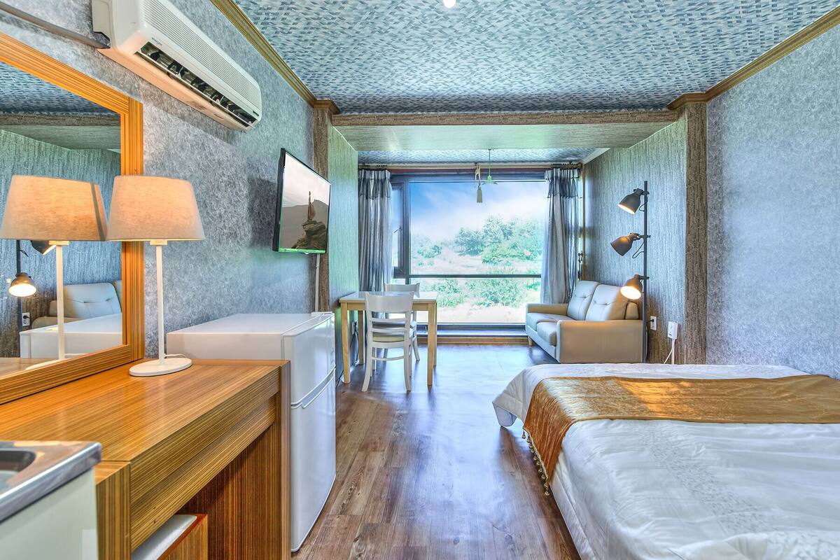 Habitación de hotel con una cama grande, un escritorio con una silla, una mesita pequeña, un espejo y un ventanal con vistas a zonas verdes.