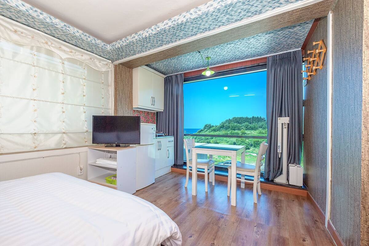 Un dormitorio con cama, televisión, una kitchenette pequeña y un balcón con vista al mar.