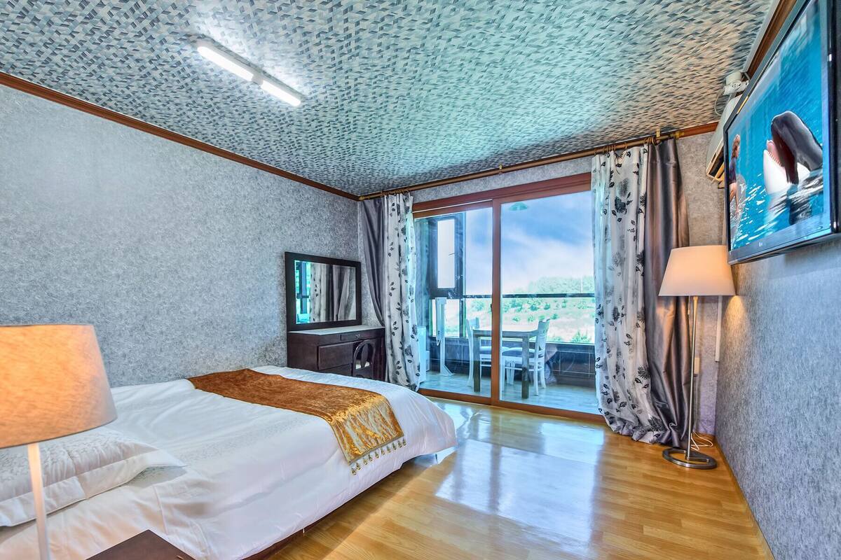 Habitación de hotel con cama, escritorio, silla, lámpara y un ventanal con cortinas.