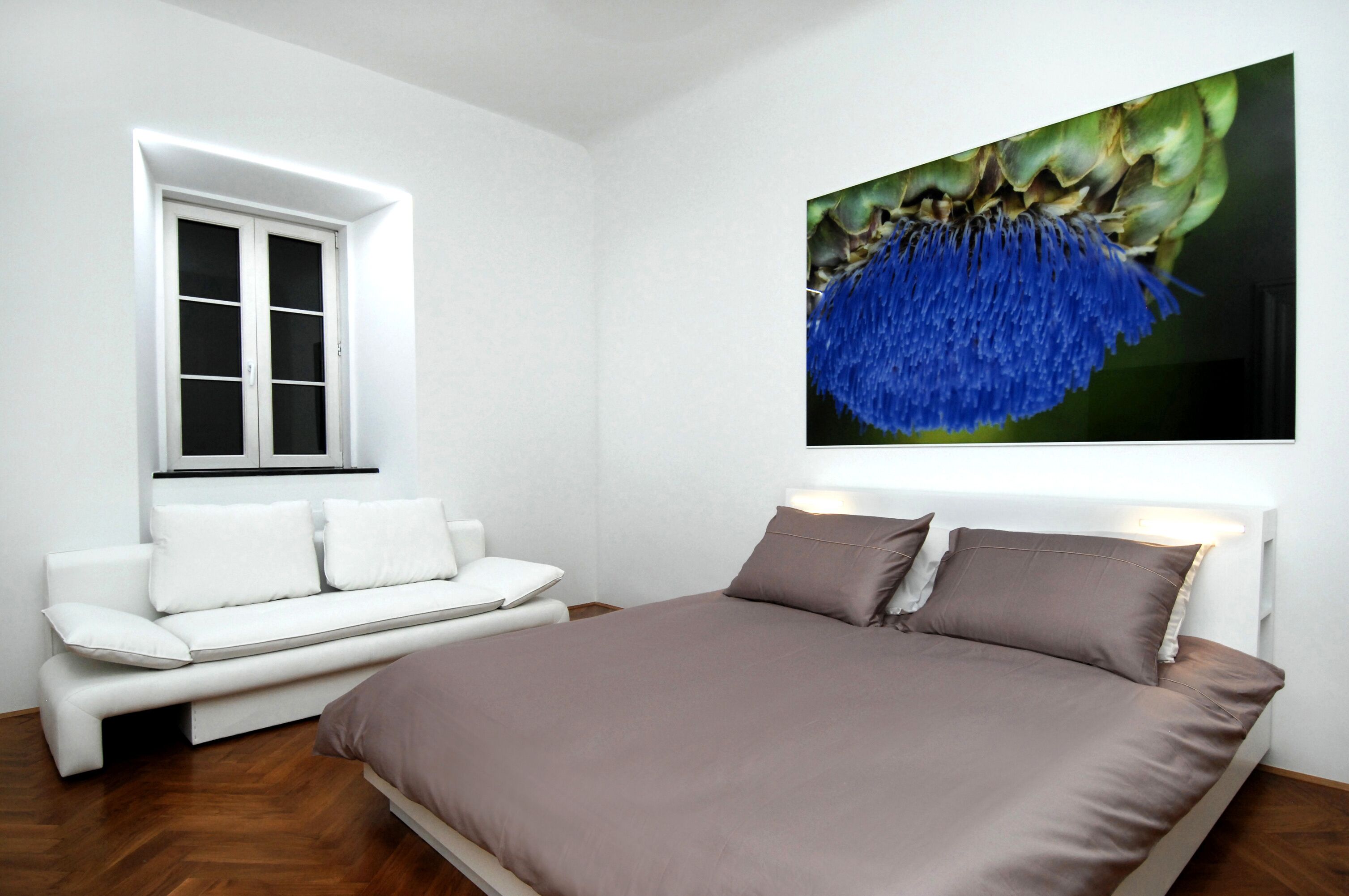A bedroom with a bed, a white sofa, a window, and a wall-mounted artwork.