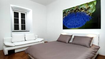 A bedroom with a bed, a white sofa, a window, and a wall-mounted artwork.