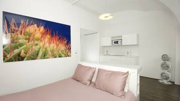 Une chambre moderne avec un lit, un grand tableau représentant un cactus, une armoire blanche et une petite kitchenette.