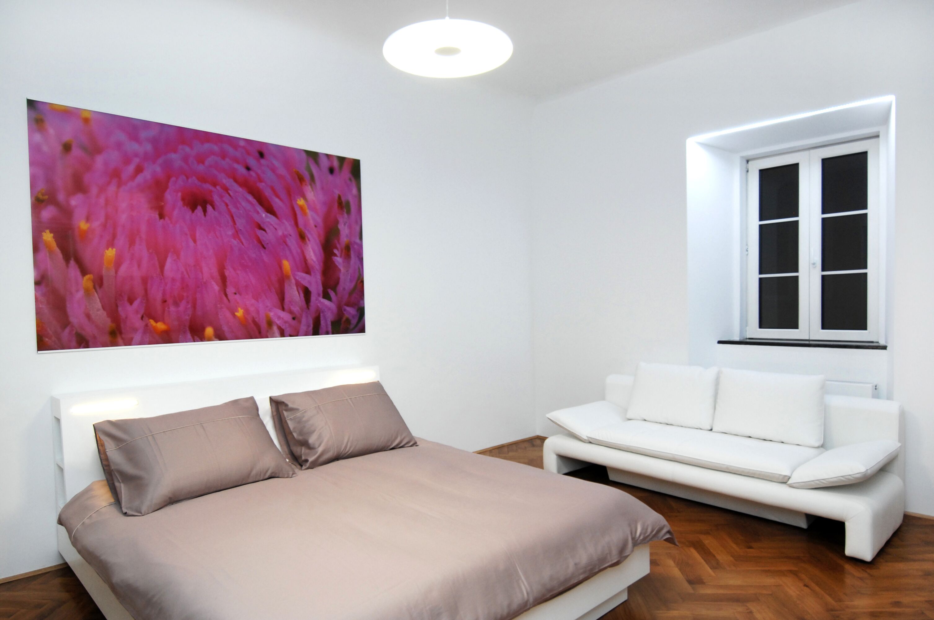 A modern bedroom with a bed, a white sofa, a large floral artwork, and a window with blinds.