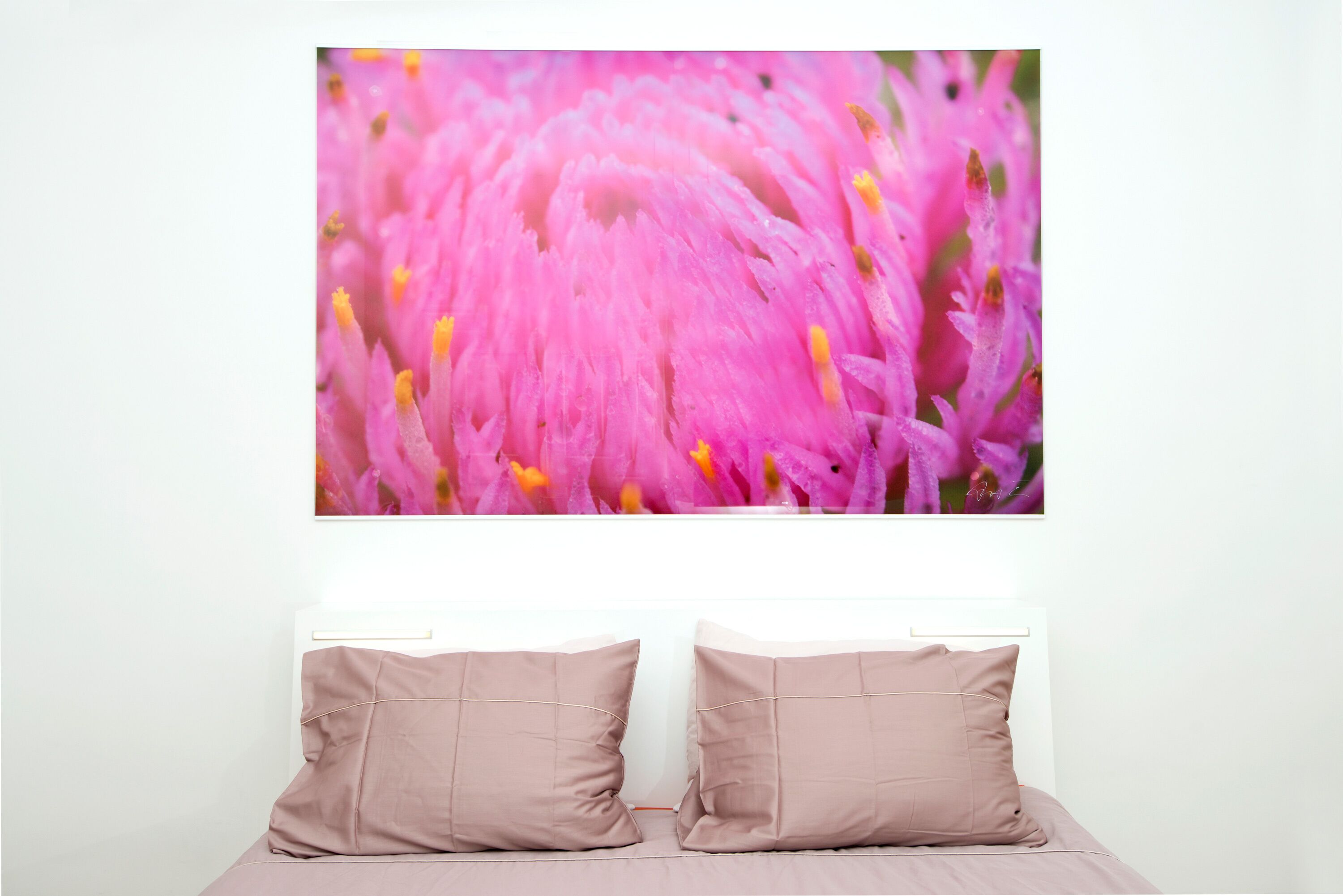 Une chambre à coucher avec un lit, deux oreillers et un grand tableau encadré représentant des fleurs roses, accroché au mur.