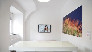 Un espace repas moderne avec une table blanche et des chaises en métal, une œuvre d’art encadrée au mur et une fenêtre avec des stores.