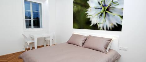 Une chambre à coucher avec un grand lit, un bureau, deux chaises, une fenêtre et un tableau représentant une fleur accroché au mur.