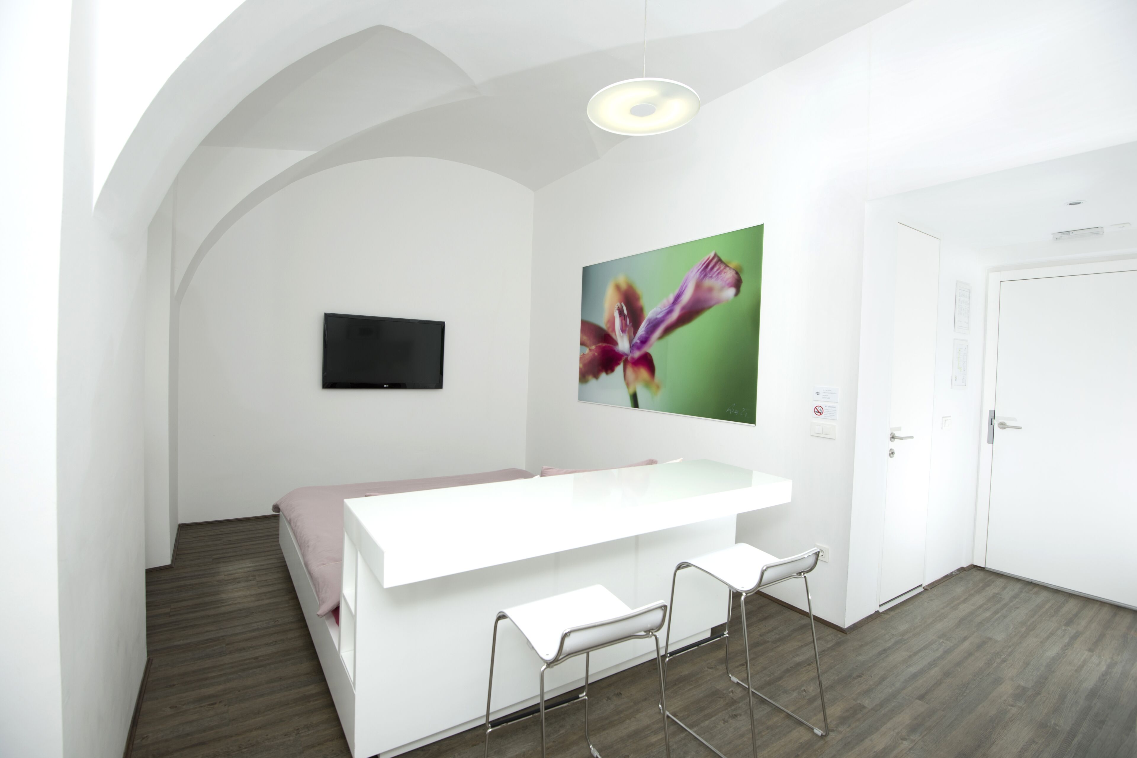 Una camera moderna con una scrivania bianca, due sgabelli da bar, un televisore a schermo piatto e un grande quadro floreale appeso al muro.