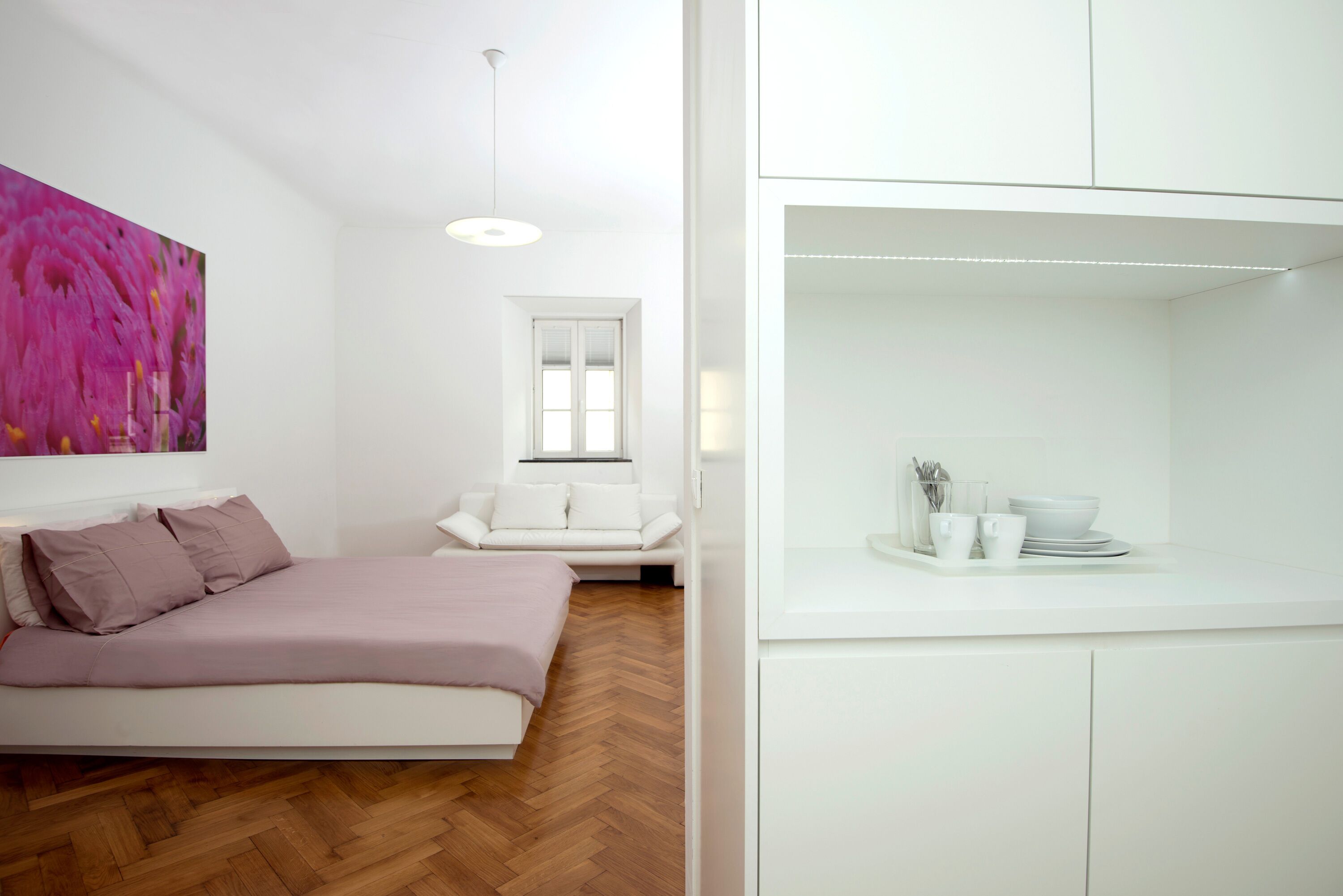 Een moderne slaapkamer met een bed, een witte bank, een groot roze bloemenkunstwerk en een witte kast met keukengerei.
