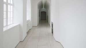 Un long couloir étroit aux murs blancs, avec de grandes fenêtres en arc de cercle d’un côté.