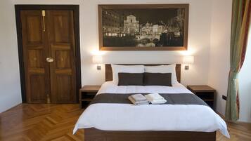 A neatly made bed with white and gray linens, two bedside tables with lamps, a wooden door, and a framed cityscape painting above the bed.