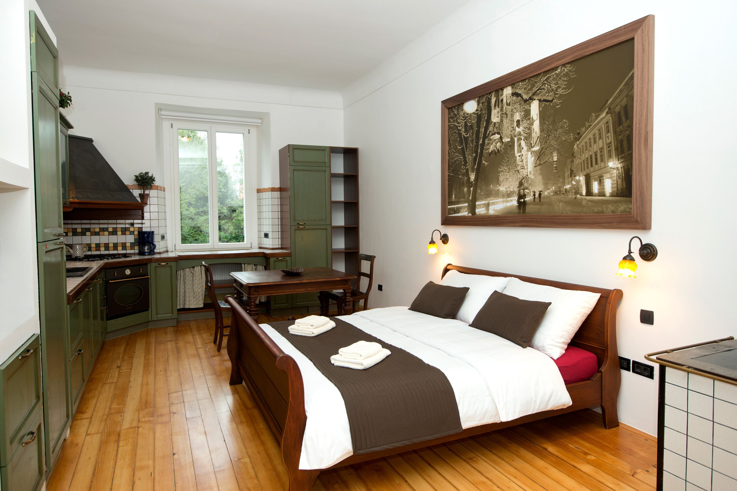 A bedroom with a wooden bed, a small desk, a kitchenette, and a large window.
