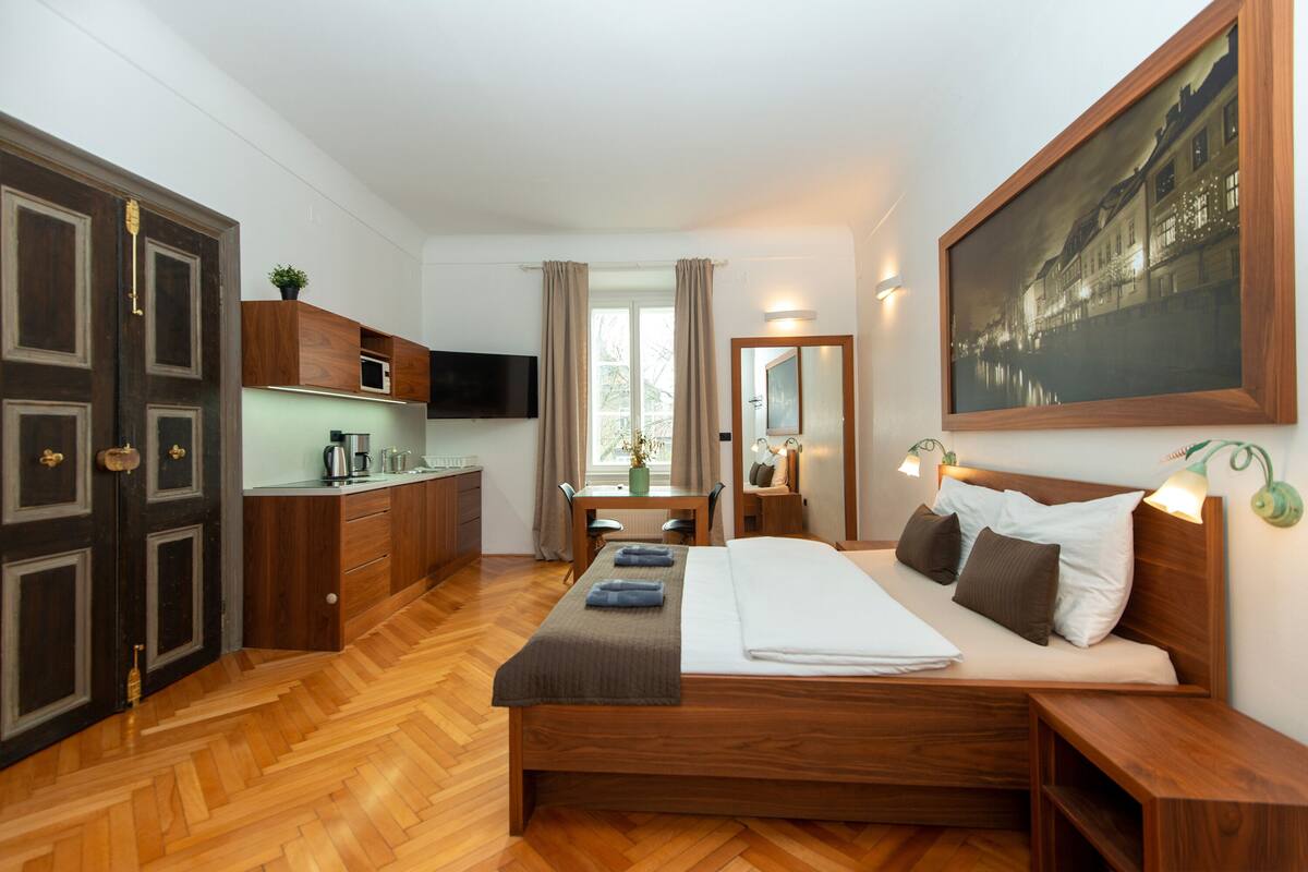 Une chambre d’hôtel comprenant un lit, des tables de chevet, un bureau avec une chaise, une télévision, un coin cuisine et une grande fenêtre avec des rideaux.