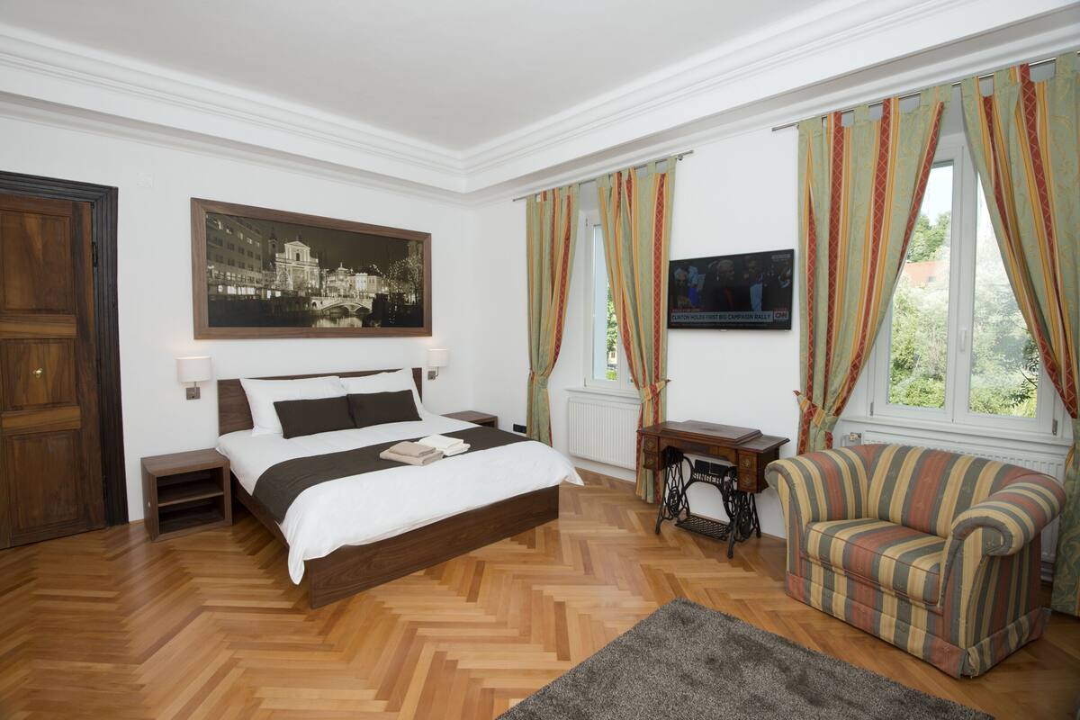 Une chambre à coucher avec un lit, un plancher en bois, un tableau, une télévision, une fenêtre avec des rideaux et un canapé.