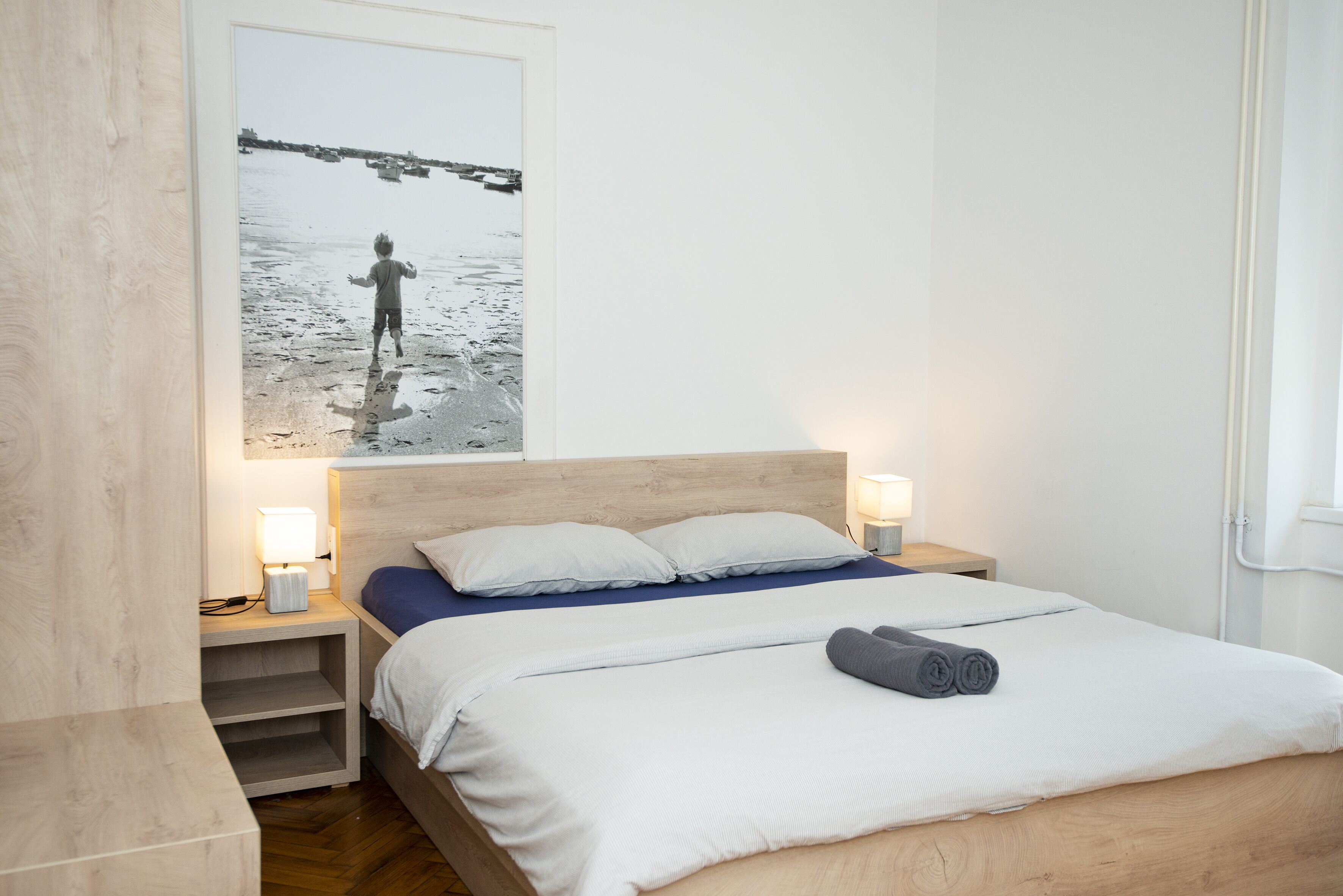 Un dormitorio con cama, dos lámparas de noche, una mesita de noche, cabecera de madera y un cuadro con una escena de playa.
