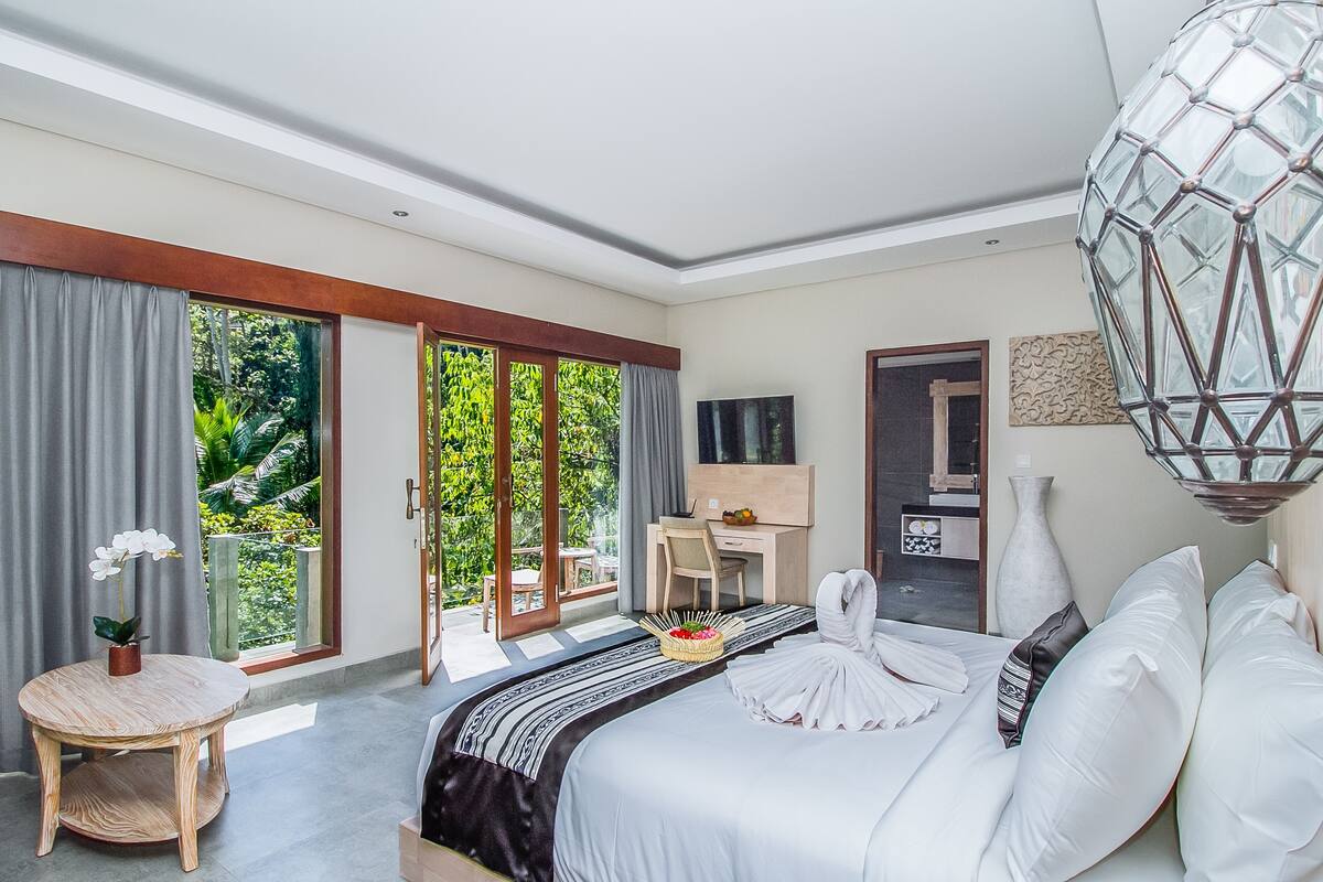 Ein Schlafzimmer mit Bett, Schreibtisch, Stuhl, rundem Tisch, einer Vase und Blick auf Grünflächen.