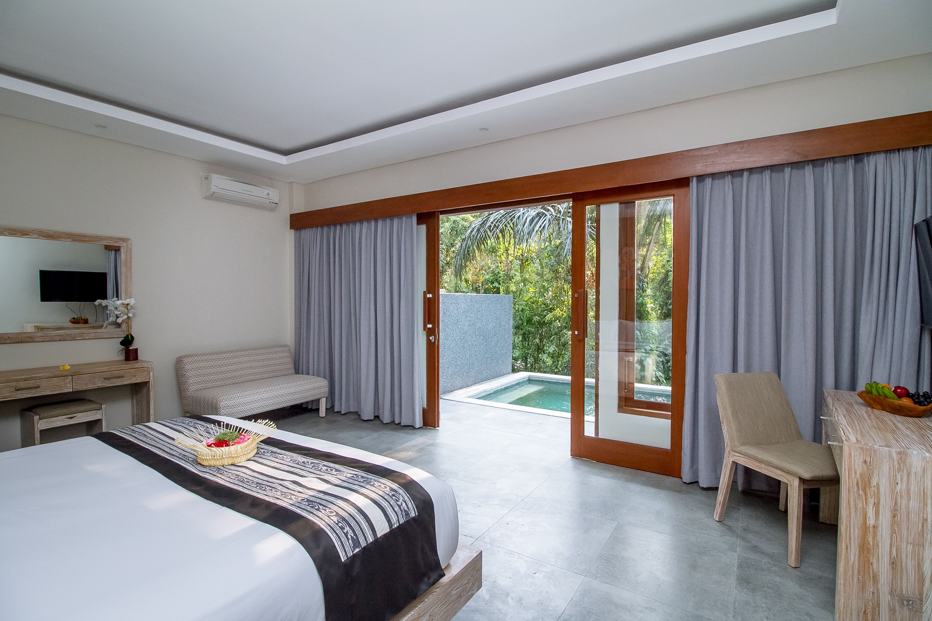 Habitación de hotel con cama, escritorio, silla, mesita con fruta y vistas a una piscina y vegetación.