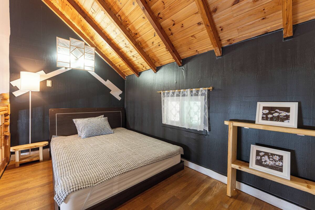 A bedroom with a wooden ceiling, a bed with a checkered bedspread, a bedside lamp, a window with a lace curtain, and a shelf with framed pictures.
