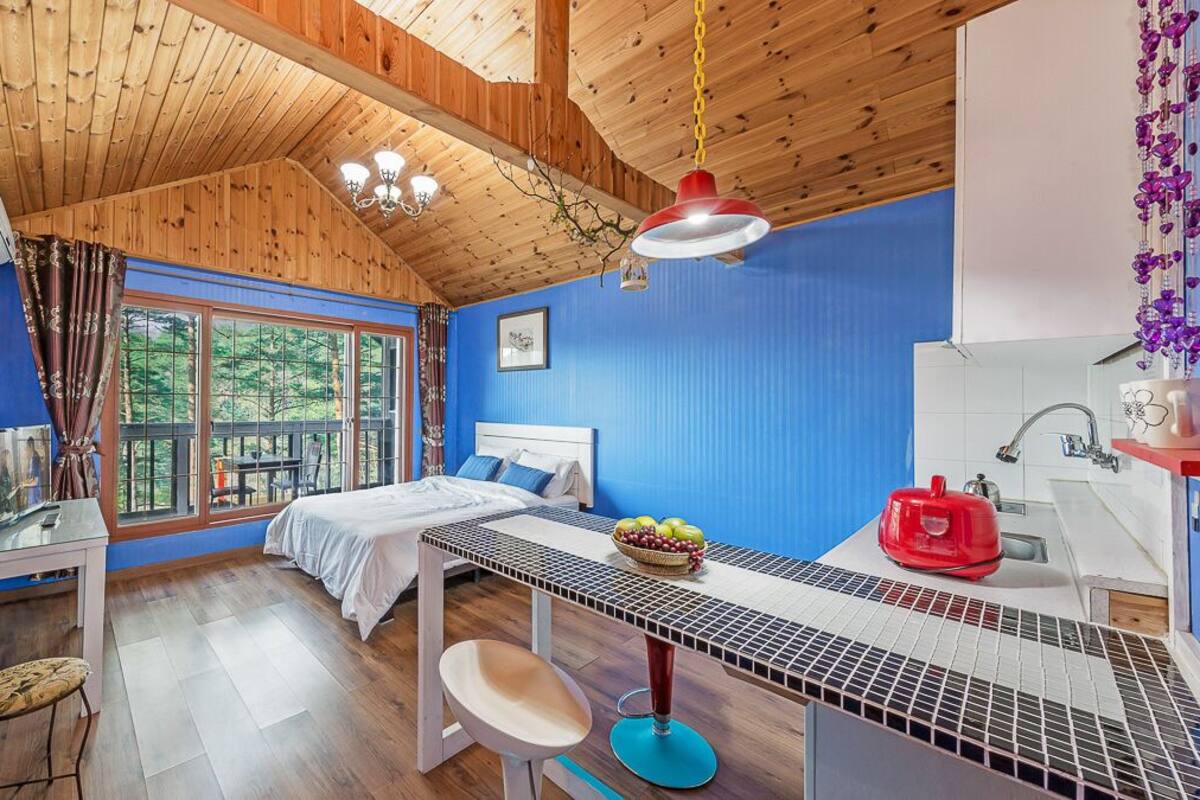 A room with a bed, a table with fruit, a sink, and a window with a view of trees.