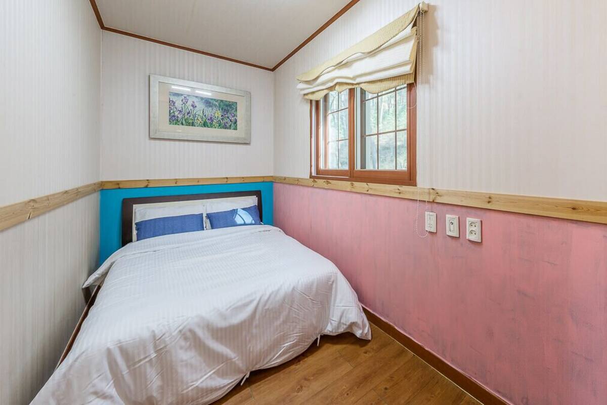 A bedroom with a bed, a window with curtains, a framed picture on the wall, and a wooden headboard.