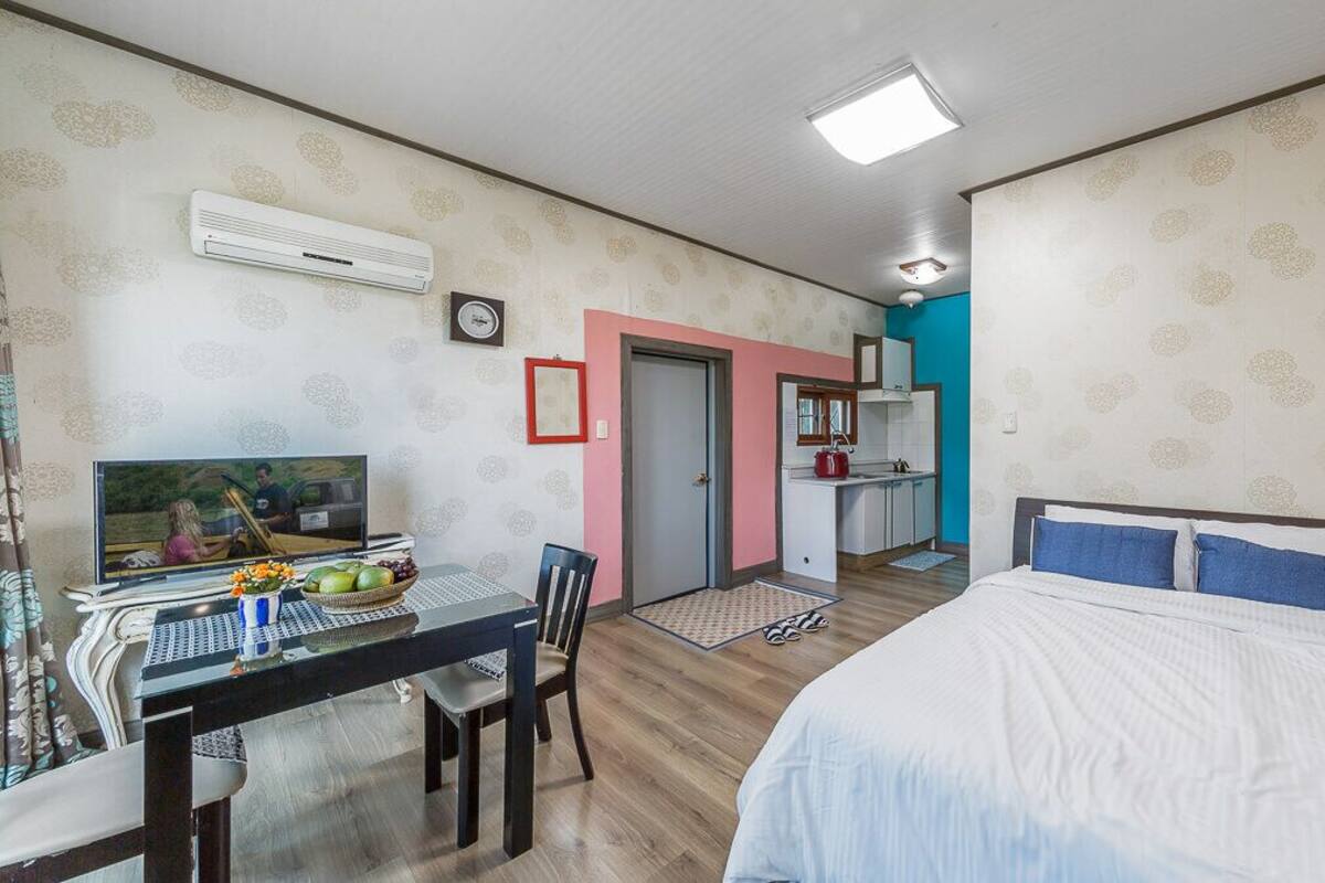A bedroom with a bed, a desk with a TV, and a kitchenette area.