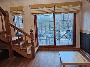 Una habitación con escalera de madera, un ventanal con vista a una terraza y mobiliario exterior, y una pequeña mesa de madera.