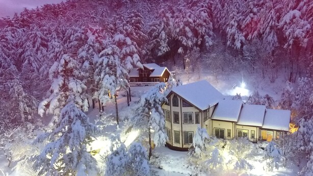 Une maison à deux étages, dont le toit est recouvert de neige, entourée d’arbres et elle-même cernée par une forêt.