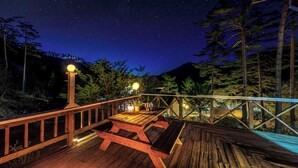 Une terrasse en bois avec une table et des chaises, entourée d’arbres et sous un ciel étoilé.