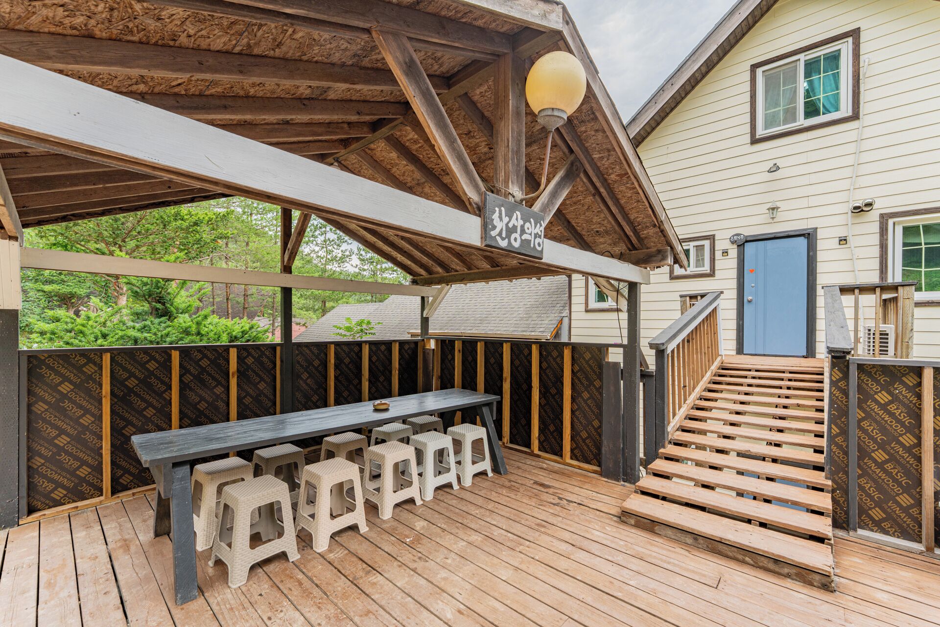 Une terrasse extérieure avec une table et des tabourets en bois, un espace salon couvert et un escalier menant à une maison.