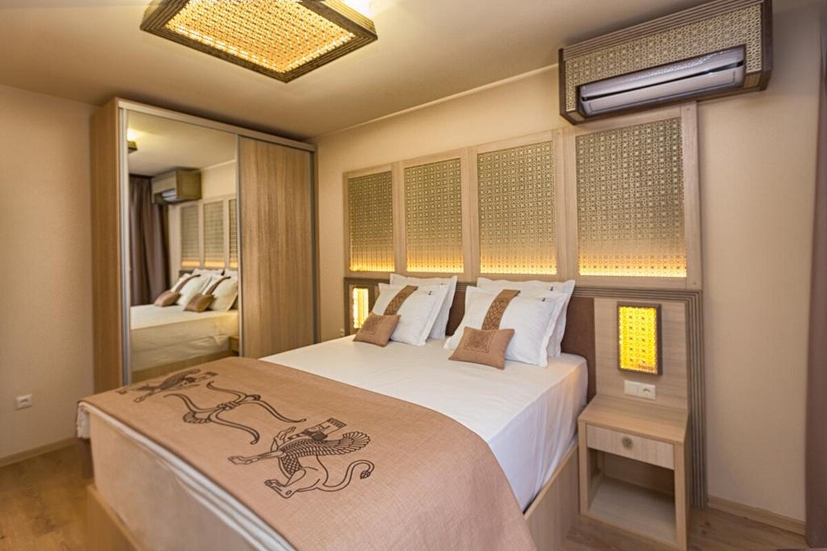 A hotel room with a bed, a bedside table, an air conditioning unit, and a mirror.