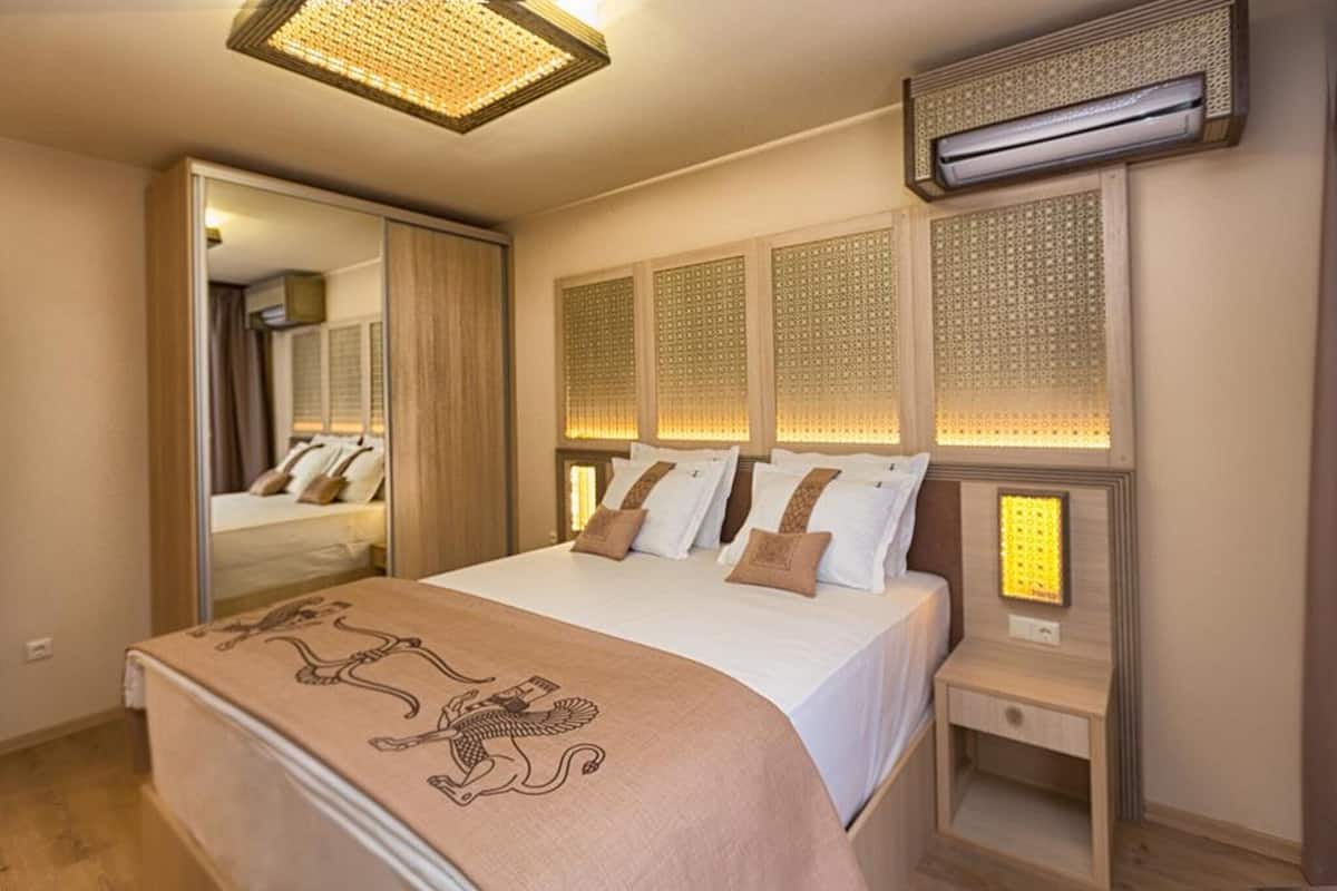 A hotel room with a bed, a bedside table, an air conditioning unit, and a mirror.