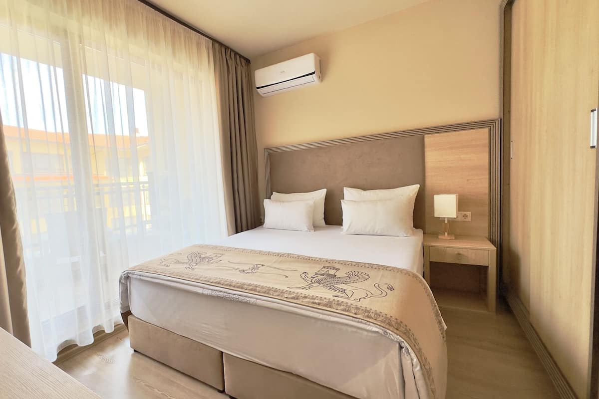 A hotel room with a bed, a bedside table with a lamp, an air conditioning unit, and a view of the outside through sheer curtains.