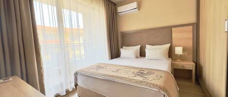 Habitación de hotel con cama, mesita de noche con lámpara, equipo de aire acondicionado y vista al exterior a través de cortinas translúcidas.