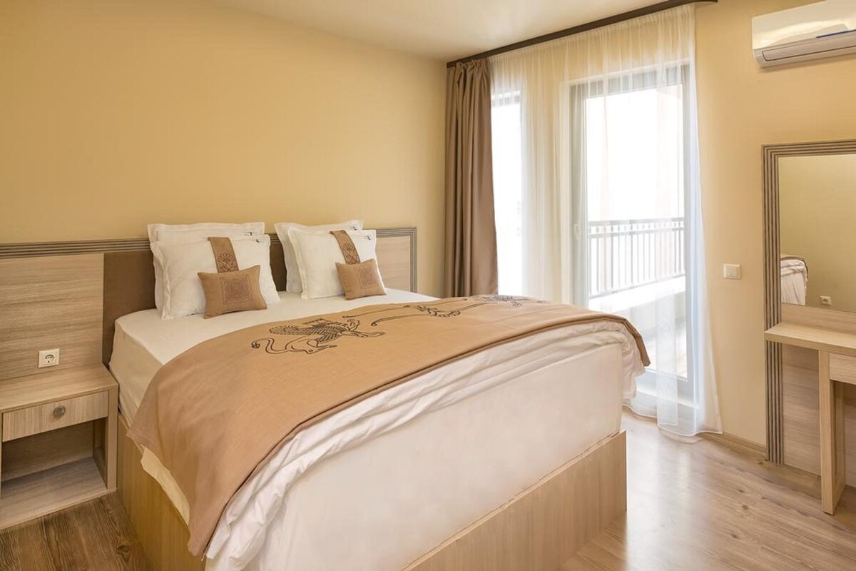 A hotel room with a bed, a bedside table, a mirror, and a window with curtains.