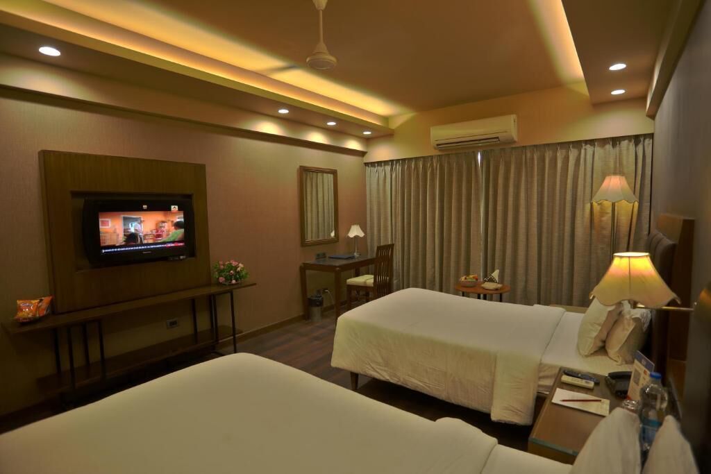 Habitación de hotel con dos camas, televisión, escritorio con silla, lámpara y ventana con cortinas.